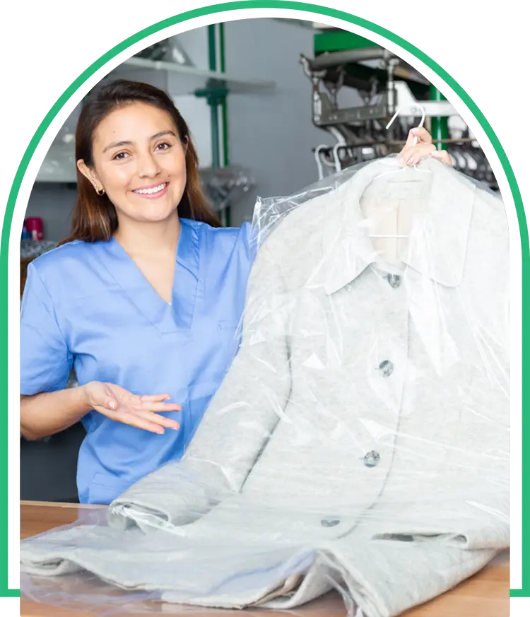 A woman in blue scrubs stands by a counter, holding up a light-colored coat covered in plastic at a dry cleaning facility.