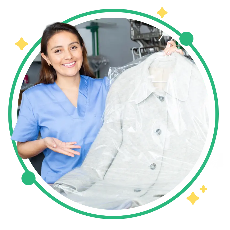 A woman in blue scrubs holds up a freshly dry-cleaned coat covered in plastic, standing in a dry-cleaning facility.