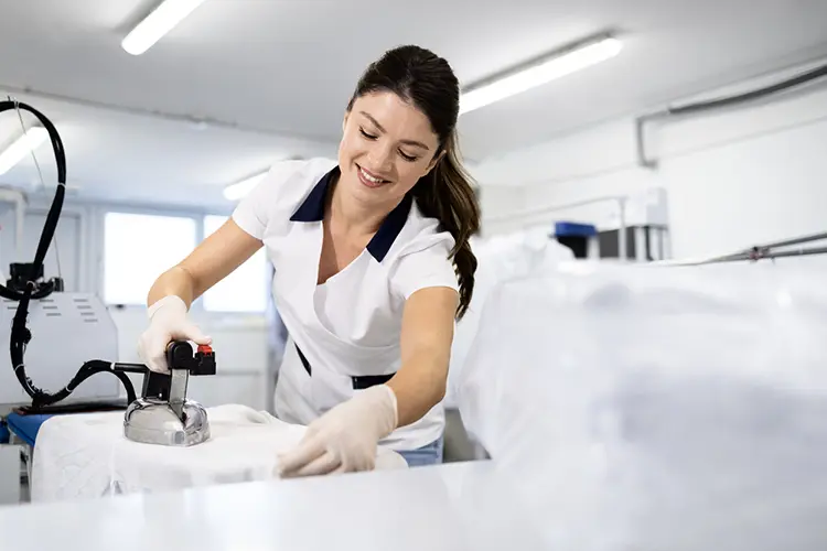 A woman in a uniform smiles while ironing clothes in a bright, modern laundry facility.