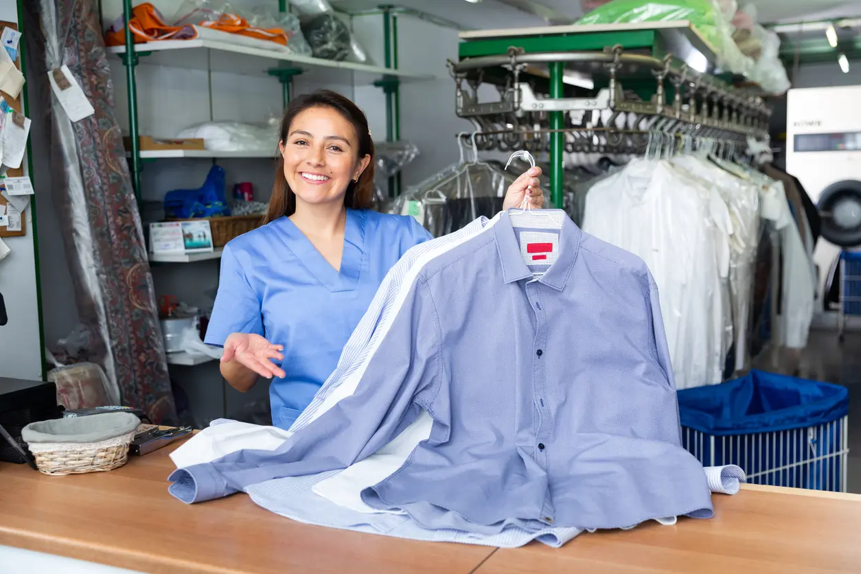 A woman in a blue uniform stands behind a counter at a dry cleaners, holding up freshly cleaned shirts on hangers. Clean clothes are visible in the background.