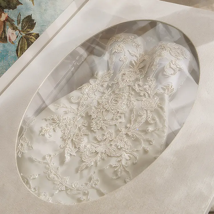 A white lace wedding dress with floral embroidery is preserved in a box with a clear oval window.