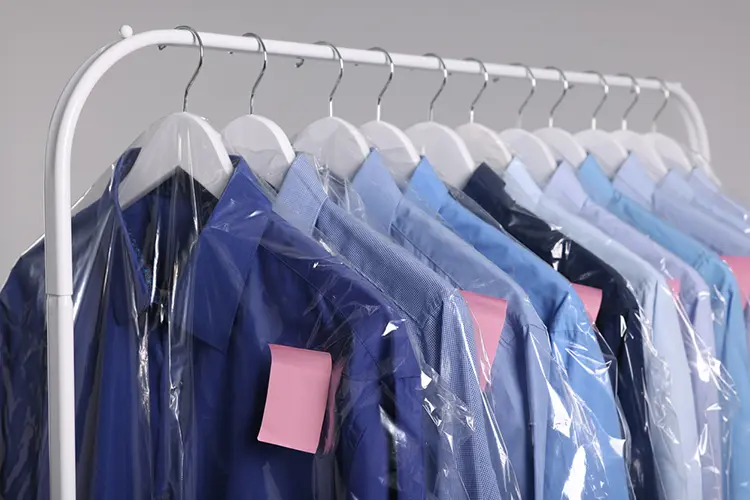 Row of blue dress shirts on hangers, covered in clear plastic garment bags, each with a pink tag attached, displayed on a white clothing rack.
