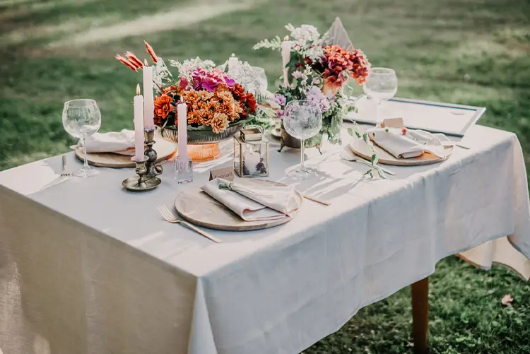 A rectangular table set outdoors with a white tablecloth, plates, wine glasses, candles, and floral centerpieces, arranged for a small gathering or meal.