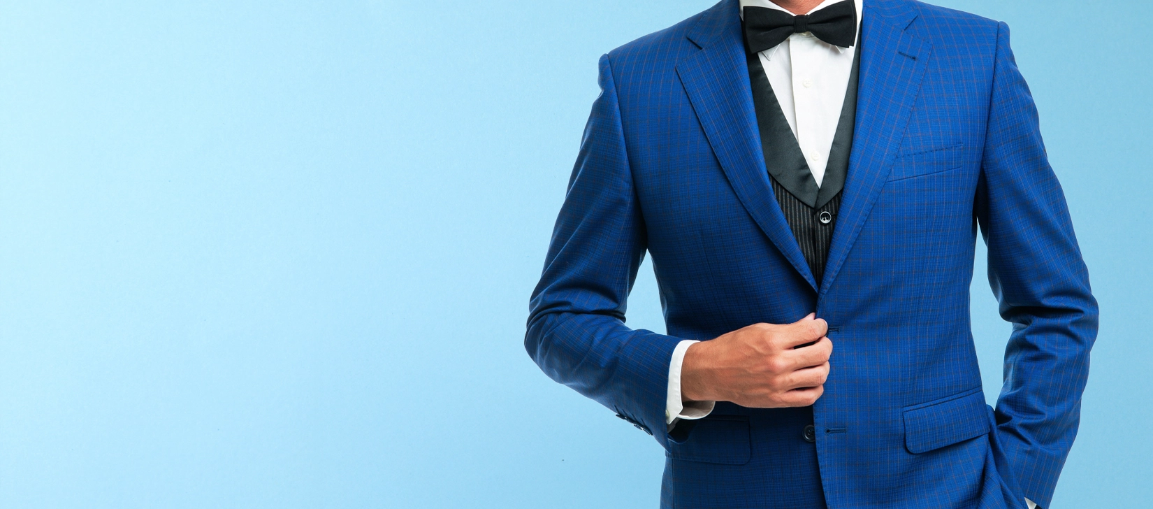 A person wearing a blue checked suit, black bow tie, and white shirt stands against a light blue background, with one hand adjusting the jacket.