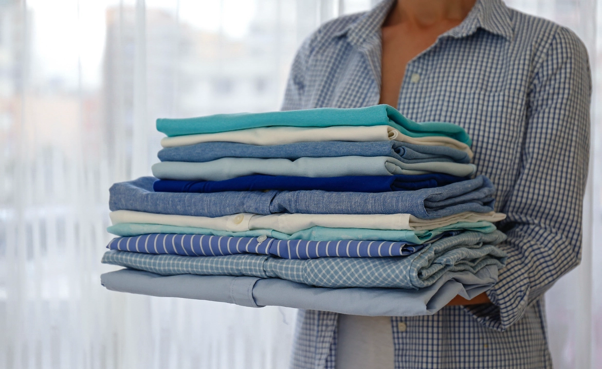 A person wearing a checked shirt holds a neat stack of folded clothes in various shades of blue and white against a light background.