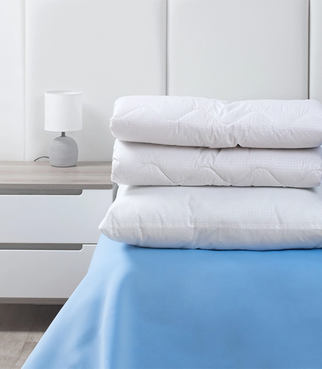 A neatly made bed with a blue sheet, two folded white comforters stacked on top, a bedside table with drawers, and a small lamp.