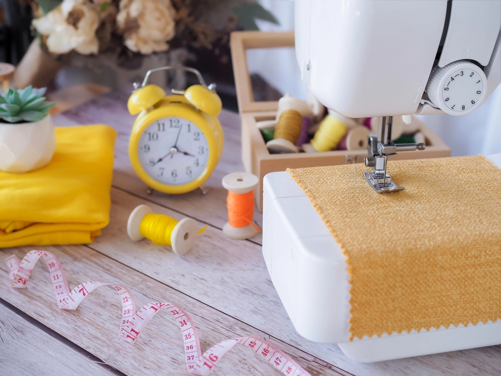 A sewing machine stitching yellow fabric on a wooden table with spools of thread, a yellow alarm clock, folded fabric, and a measuring tape nearby.