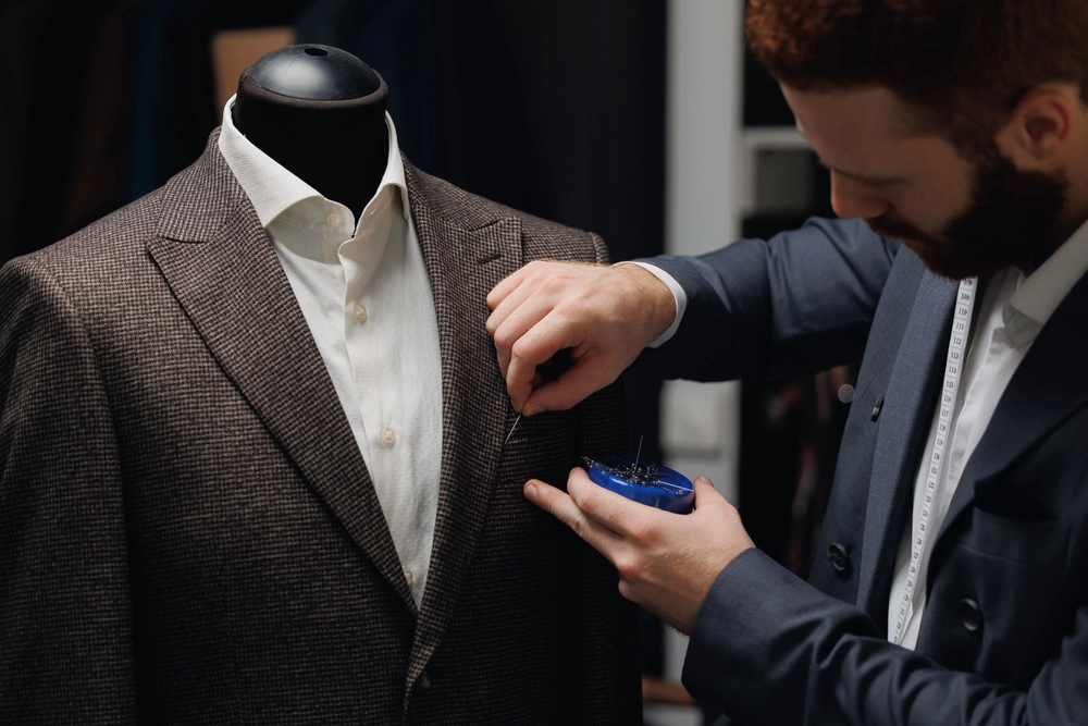 A tailor adjusts the lapel of a brown checked suit jacket on a mannequin using pins, with a measuring tape draped around his neck.