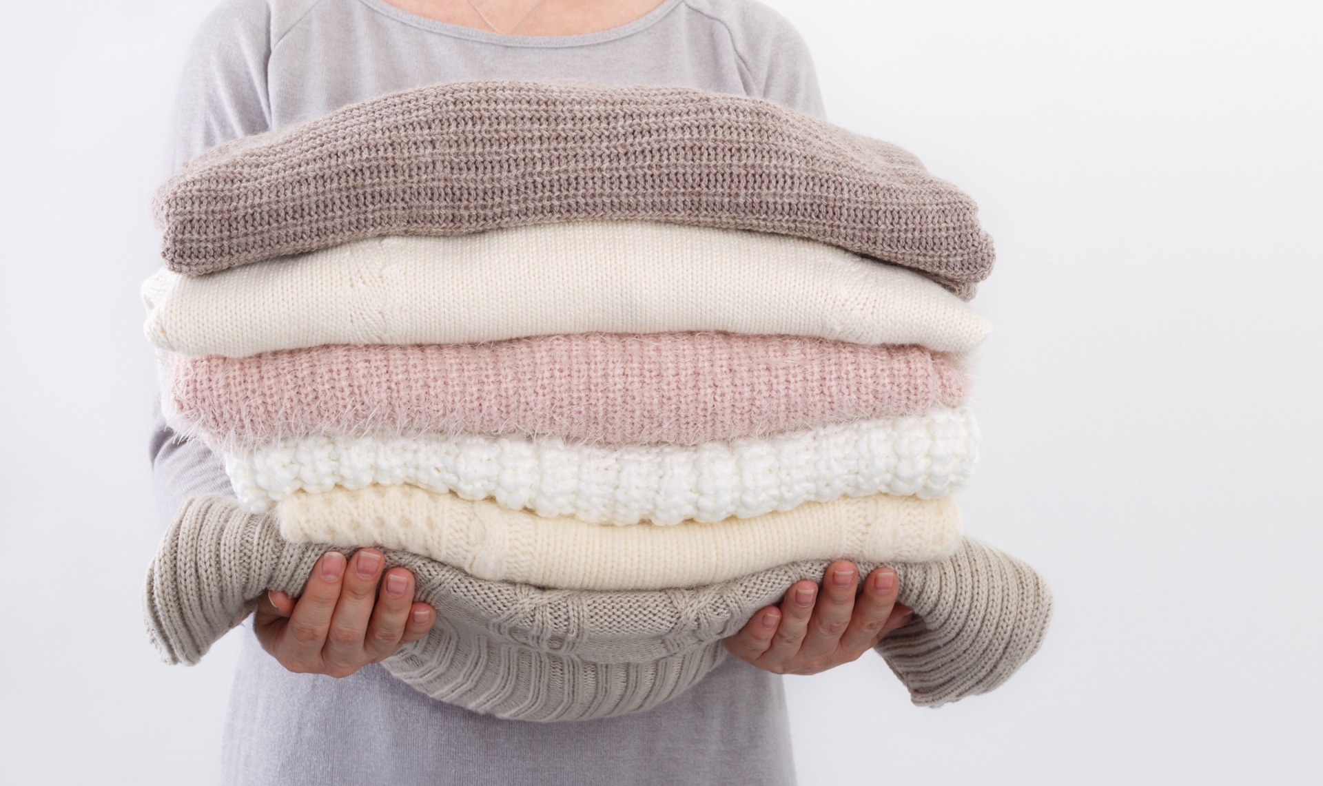 A person holds a stack of neatly folded sweaters in neutral colors, including beige, pink, cream, and white, against a plain background.