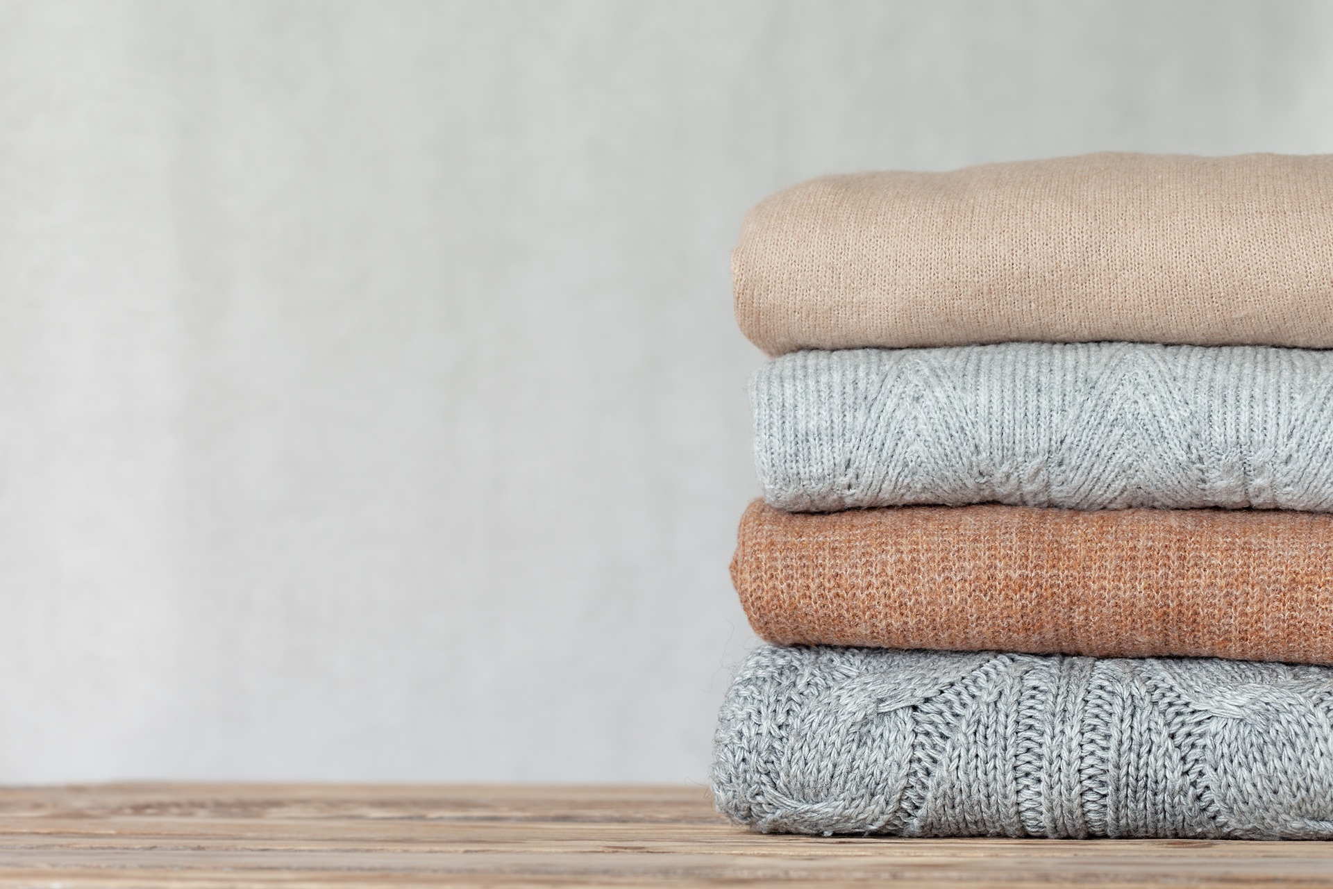 A stack of four folded sweaters in neutral colors sits on a wooden surface against a plain, light background.