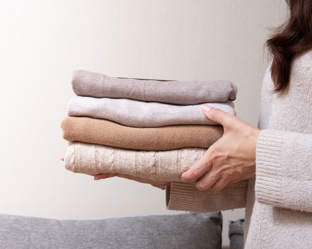 A person holding a neatly folded stack of sweaters in neutral colors against a plain background.