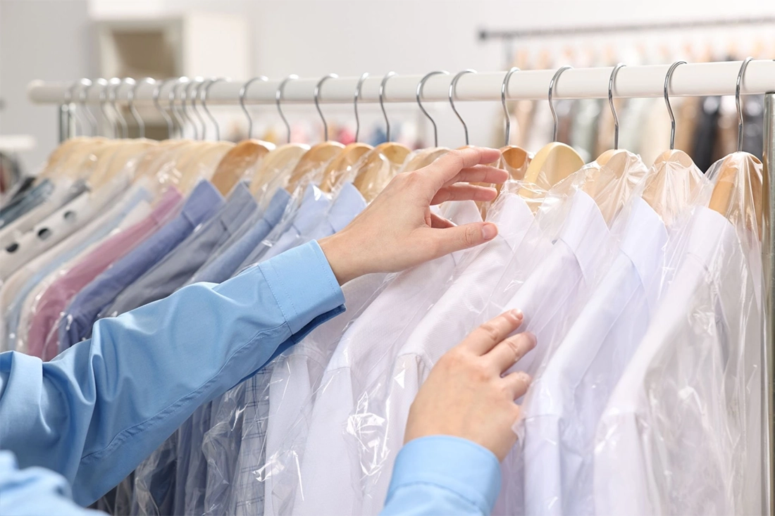 Person's hands sorting through freshly dry-cleaned dress shirts covered in plastic garment bags, hanging on a clothing rack.