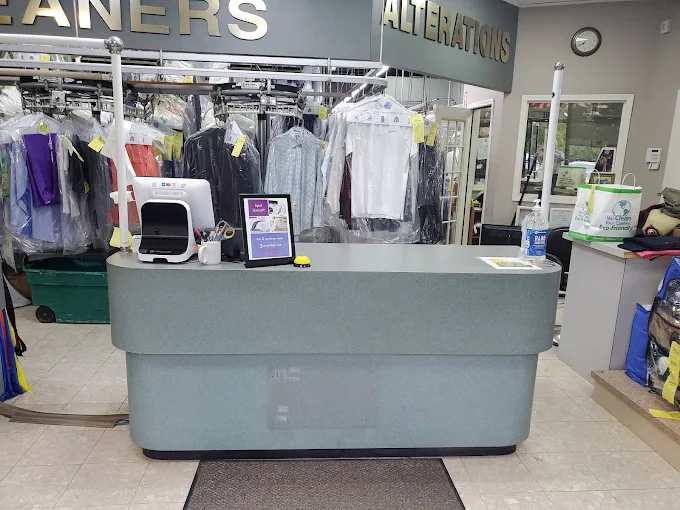 A dry cleaning shop counter with a point-of-sale system, cleaning tags, a tip jar, and hanging clothes in the background.