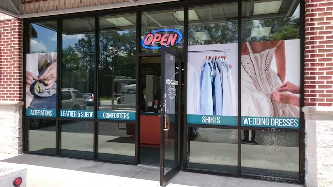 Storefront with large windows displaying images and text for alterations, leather & suede, comforters, shirts, and wedding dresses; door is open and a lit "OPEN" sign is visible.