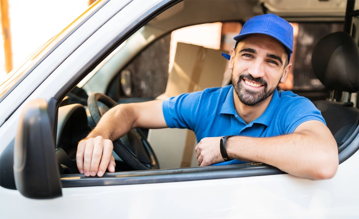 A delivery driver wearing a blue polo shirt and cap smiles while sitting in the driver’s seat of a white vehicle with packages in the back.