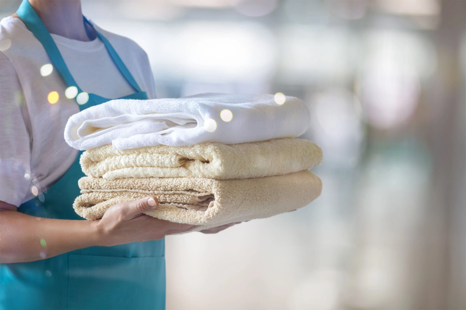 A person in a blue apron holds a stack of clean folded towels, with a blurred background.