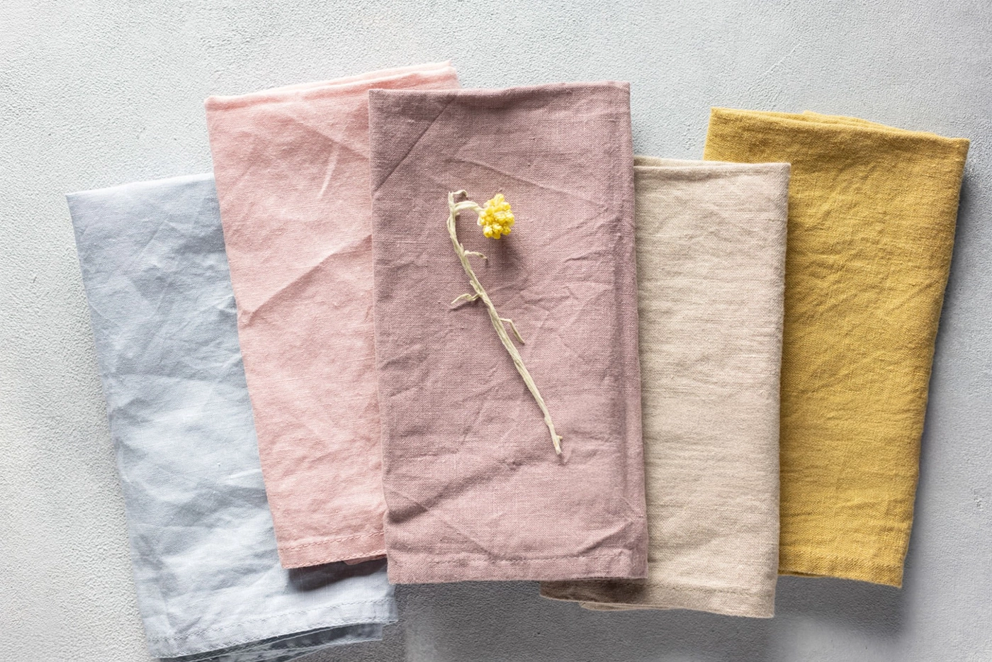 Five folded linen napkins in pastel colors—blue, pink, mauve, beige, and yellow—are arranged side by side on a light surface, with a dried flower on the mauve napkin.