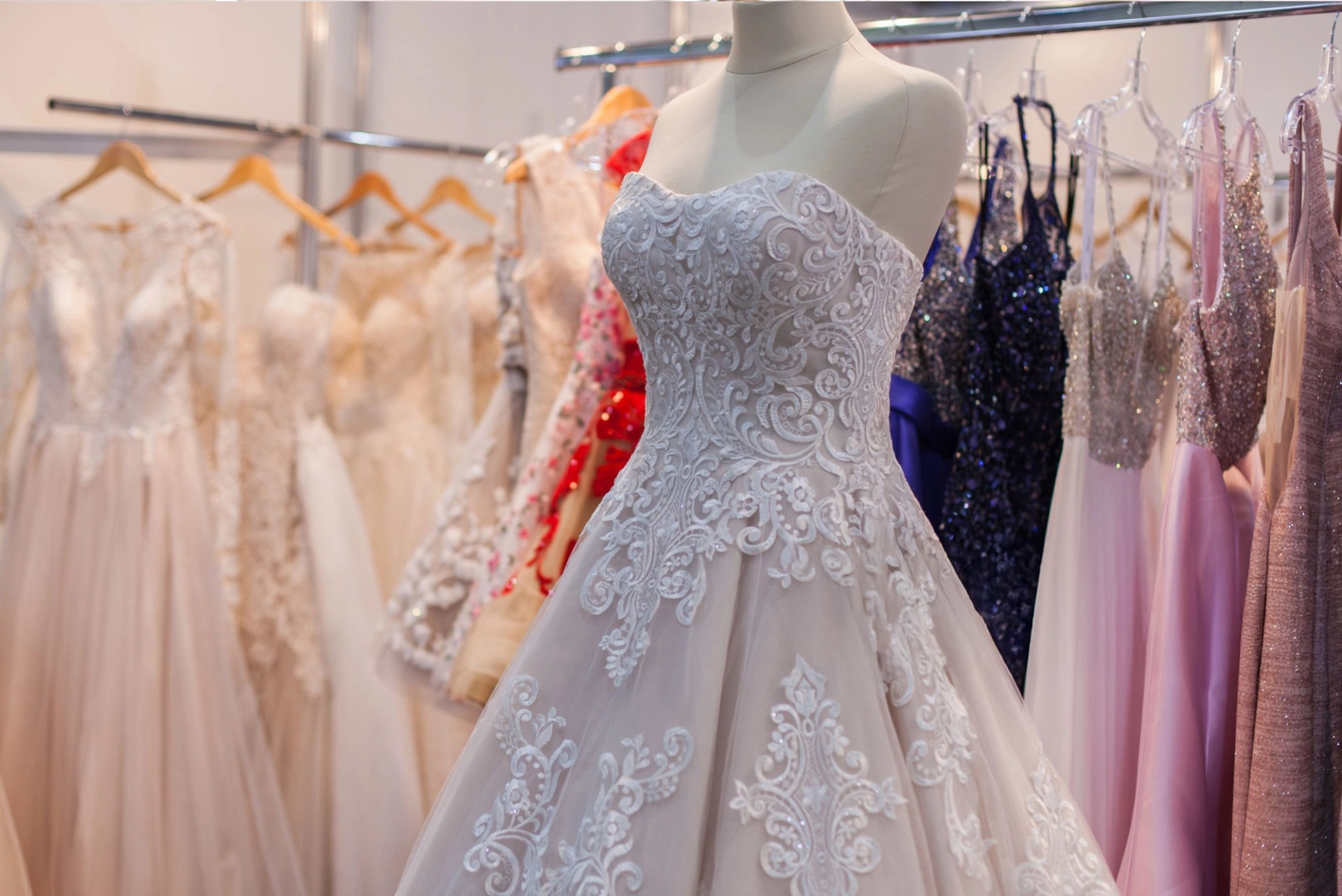 A white mannequin displays a detailed white lace wedding dress, with various formal dresses hanging on racks in the background.