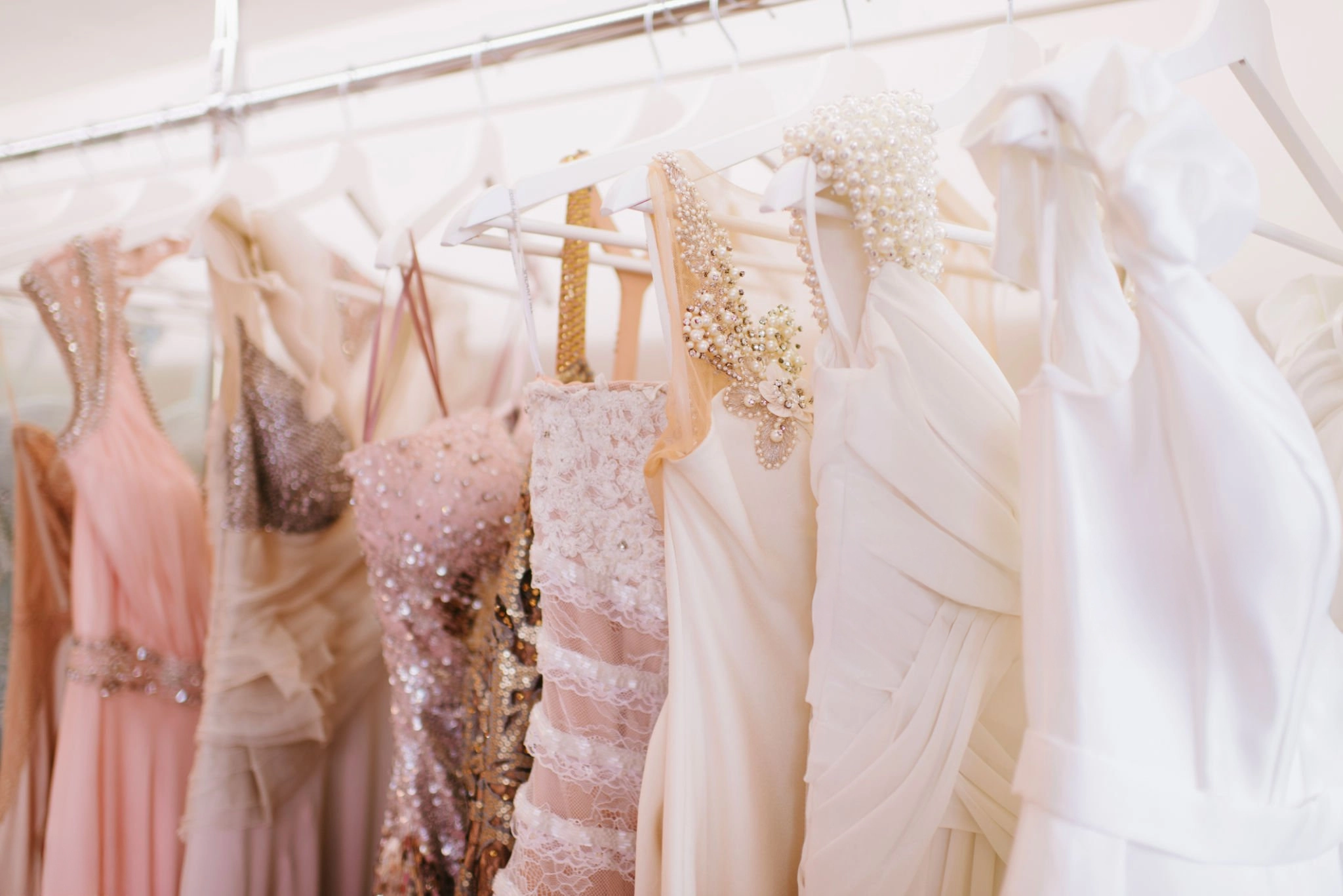 A row of elegant light-colored dresses, including embellished and lace gowns, hanging on white hangers in a closet or boutique.