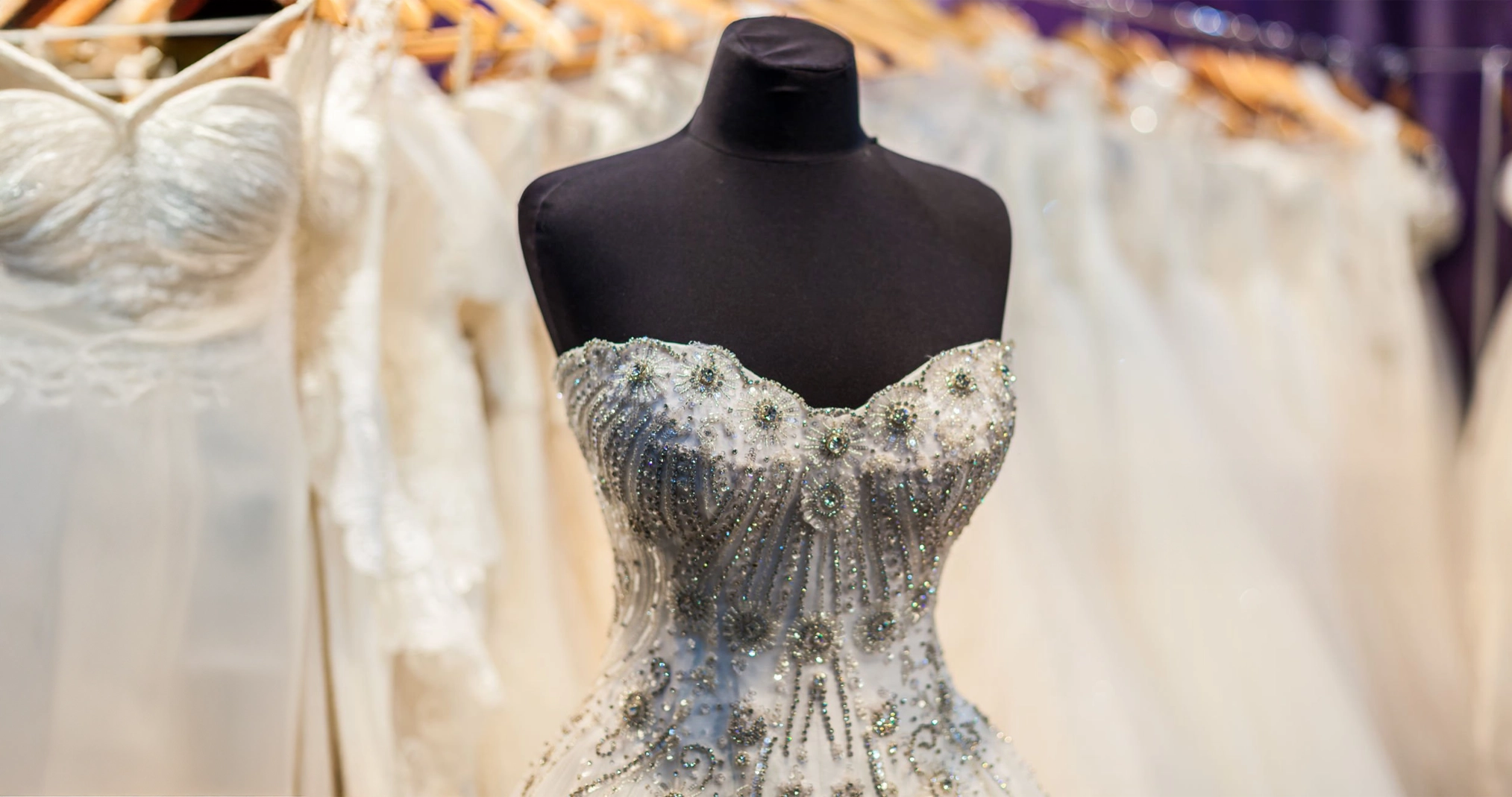 A beaded, strapless wedding dress on a black mannequin stands in front of a rack of other white wedding dresses on hangers.